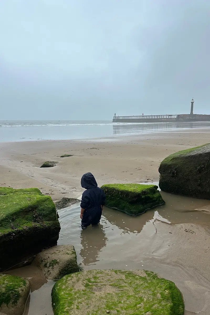 A small boy stands on a beach in a rock pool, up to his knees in water. He wears a blue puddle suit and wellies. The sky is cloudy and it is raining. In the background there is a pier with a lighthouse.