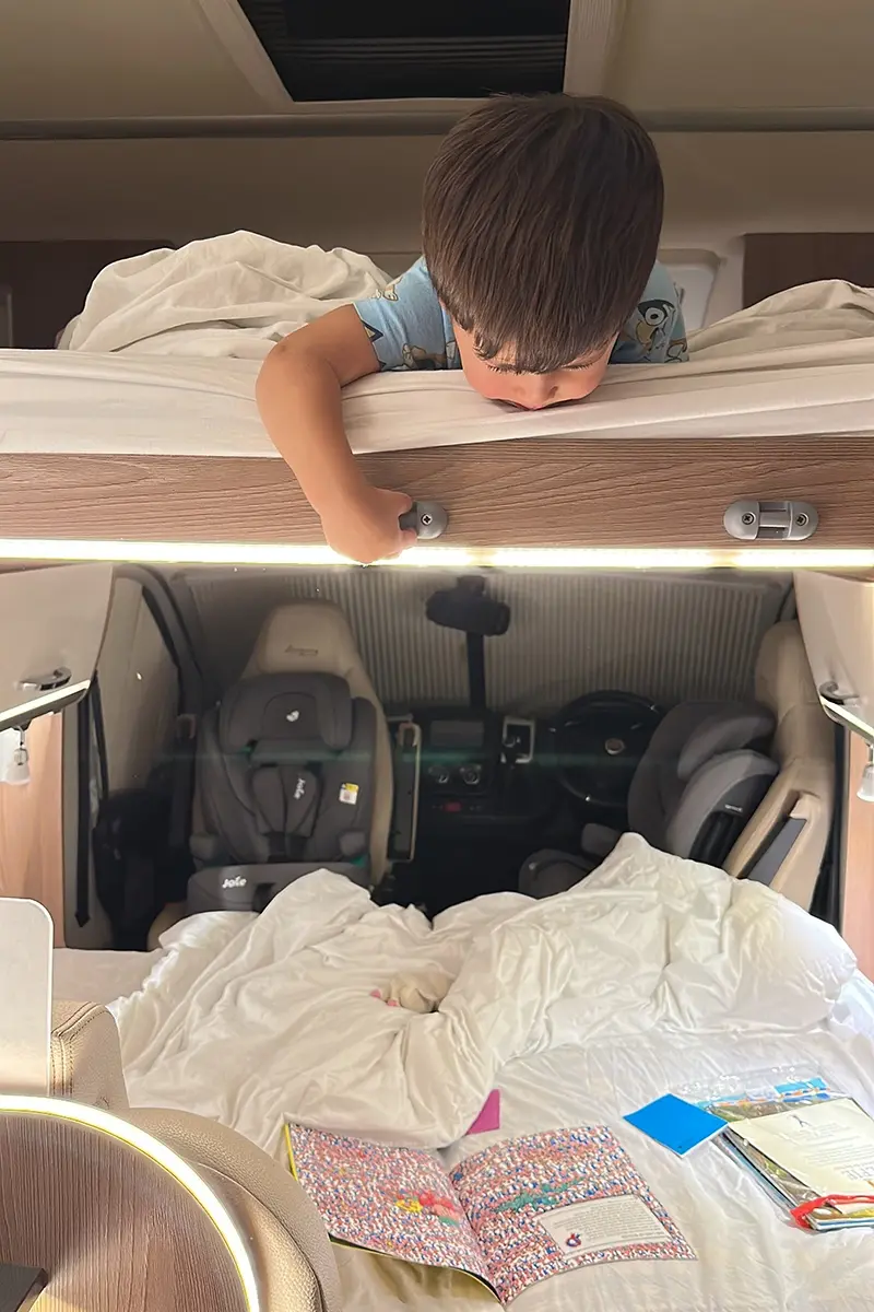 A small boy lies on a drop-down bed inside a motorhome. He is looking down to the cabin below where there is another bed and a bedtime story open on the bed. It is night time.