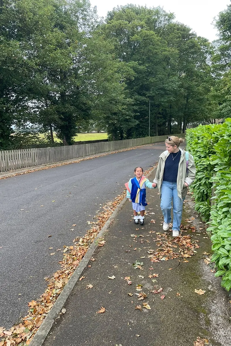 A mother and daughter walk on a pavement to school. The street is quiet and surrounded by trees. The girl wears a blue cardigan and a checkered blue school summer dress, white socks, and black shoes. She carries a book bag and has brown hair. The mother wears jeans, a blue jumper, and a green raincoat. She has blonde hair.