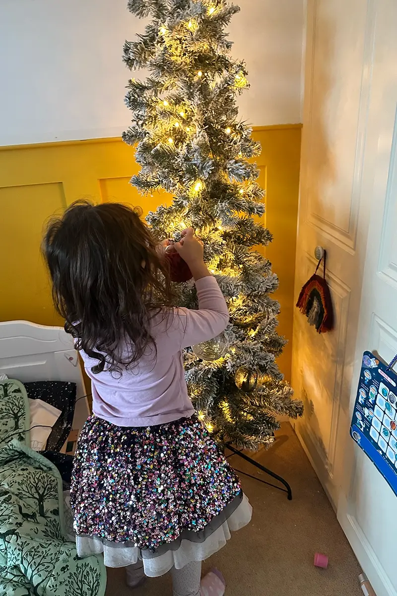 A little girl wearing a sparkly sequin skirt puts a bauble on a Christmas tree covered in twinkly fairy lights