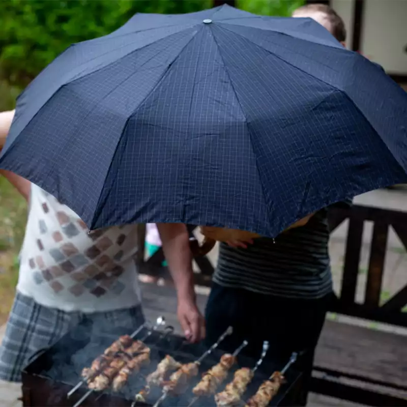 men barbecuing in the rain holding a blue umbrella